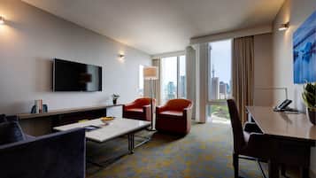 Een hotelkamer met een flatscreen televisie, twee rode stoelen, een eettafel, een salontafel en een groot raam met uitzicht op de stad.