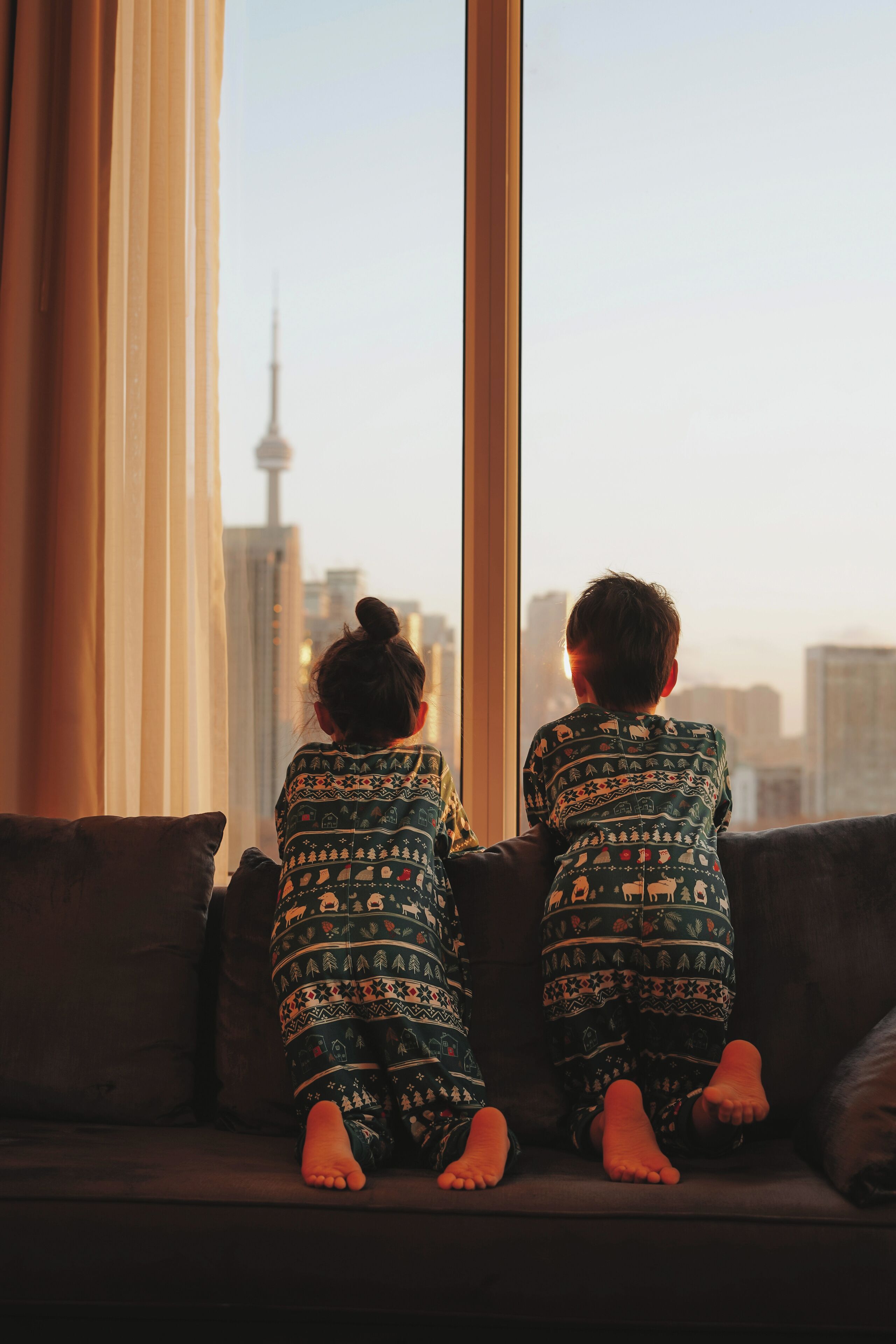 Two children sitting on a couch facing a window with a cityscape view.