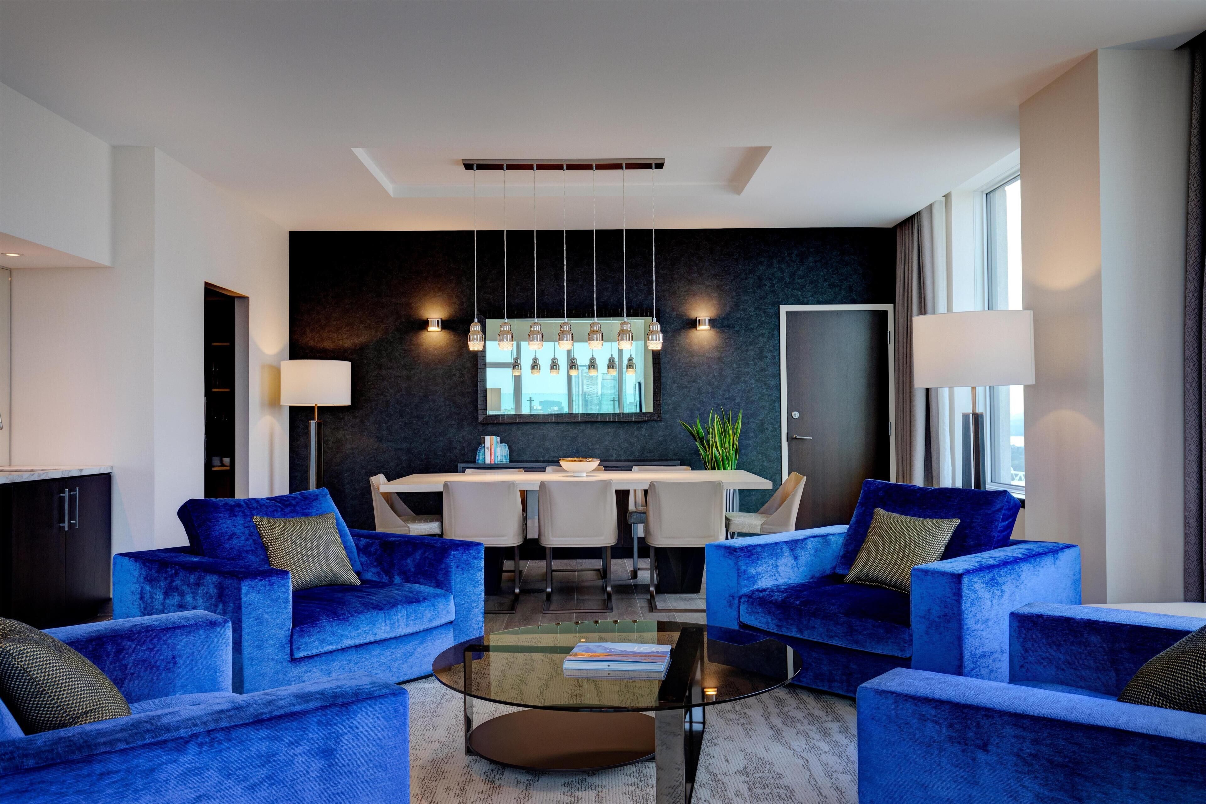 A modern living room with blue sofas, a glass coffee table, and a dining area with a white table and chairs.