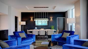 A modern living room with blue sofas, a glass coffee table, and a dining area with a white table and chairs.