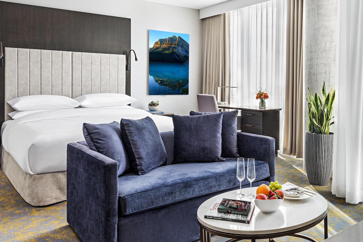 A hotel room with a bed, a sofa, a desk, a chair, a small table with a bowl of fruit and two glasses, and a large window with curtains.