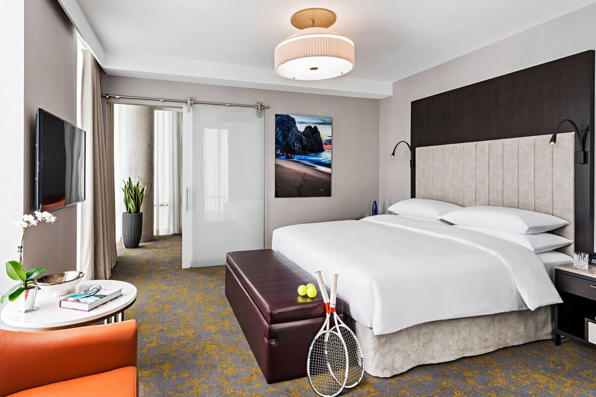 A modern hotel room with a large bed, a flat-screen TV, a small round table with a plant, a bench, and tennis rackets.