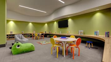 A children's library with colorful chairs, a green bean-shaped bean bag, and a flat-screen TV mounted on the wall.