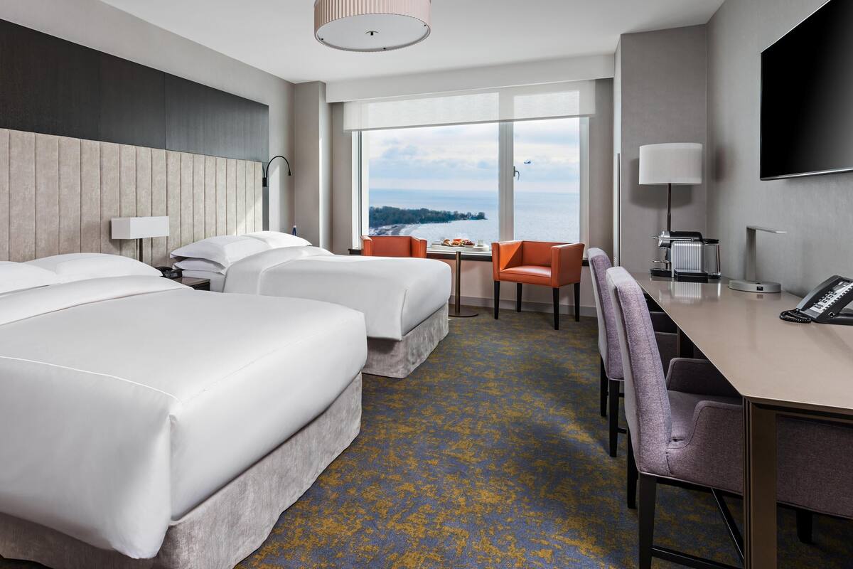A hotel room with two beds, a desk, a chair, a TV, and a view of the sea.