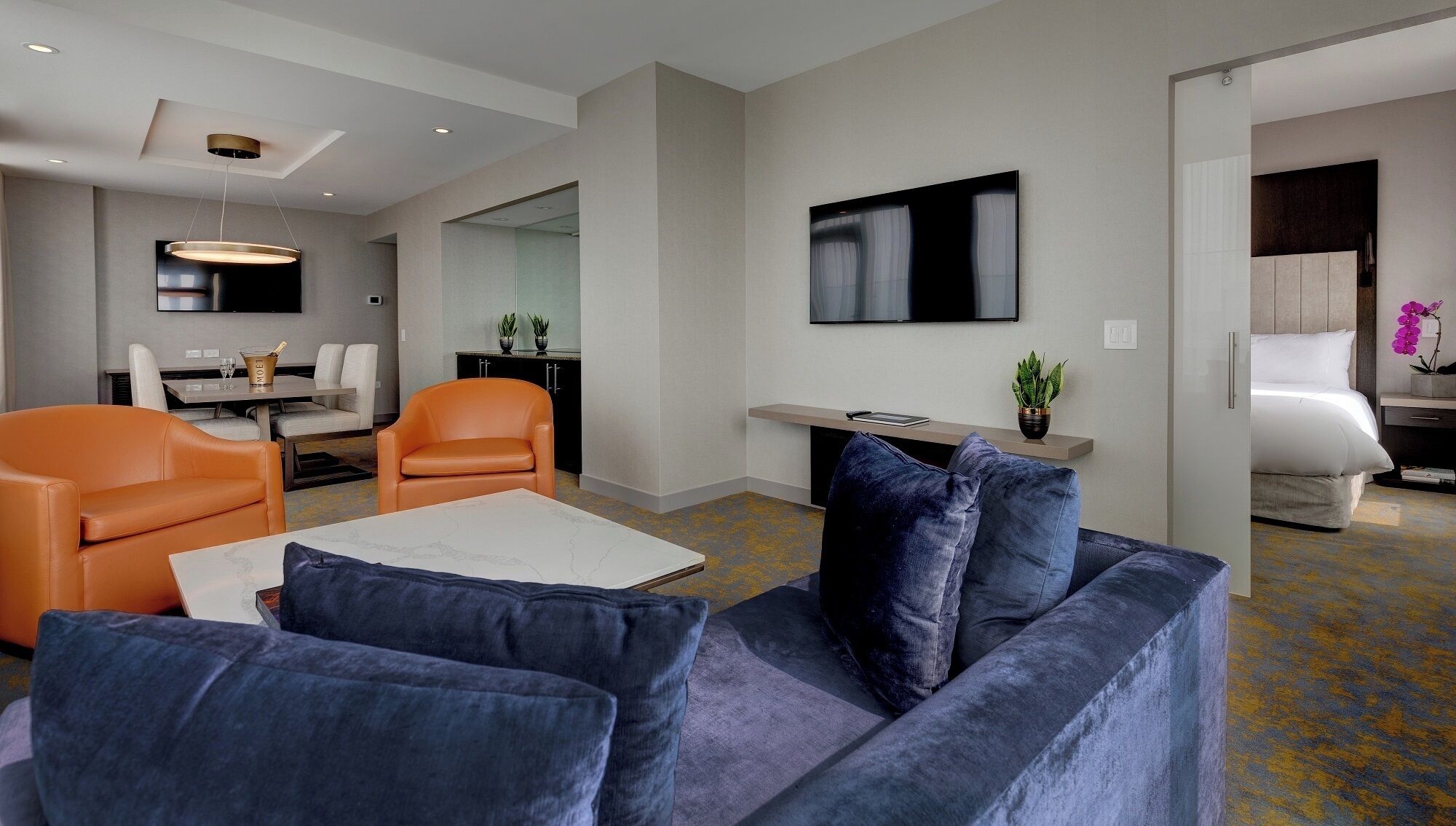 A modern living room with a sofa, armchairs, a coffee table, and a TV. There is a dining area with a table and chairs, and a kitchen area with a fridge and cabinets.