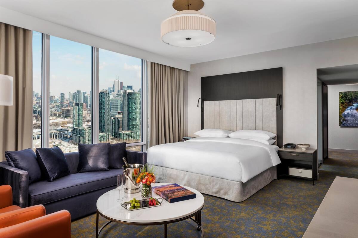A hotel room with a large bed, a sofa, a small table with a book and flowers, and a city view.