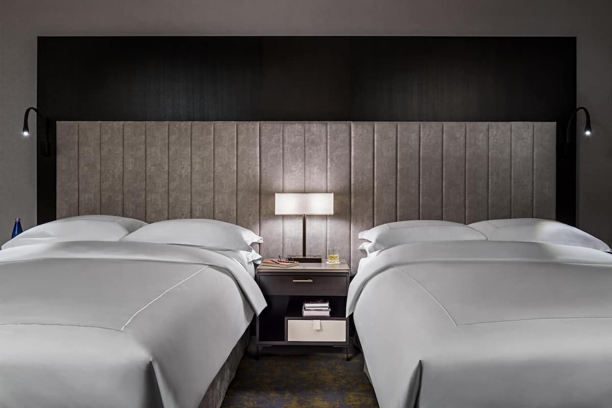 A hotel room with two beds, a nightstand with a lamp, and a small drawer.