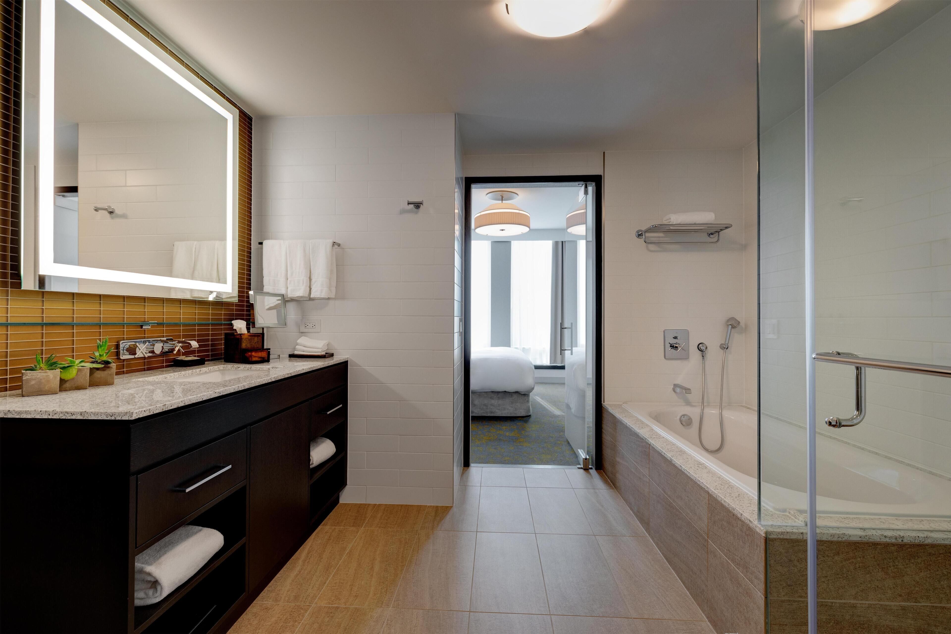 Een moderne badkamer met een grote spiegel, een douchecabine van glas en een zichtbare slaapkamer door een geopende deur.