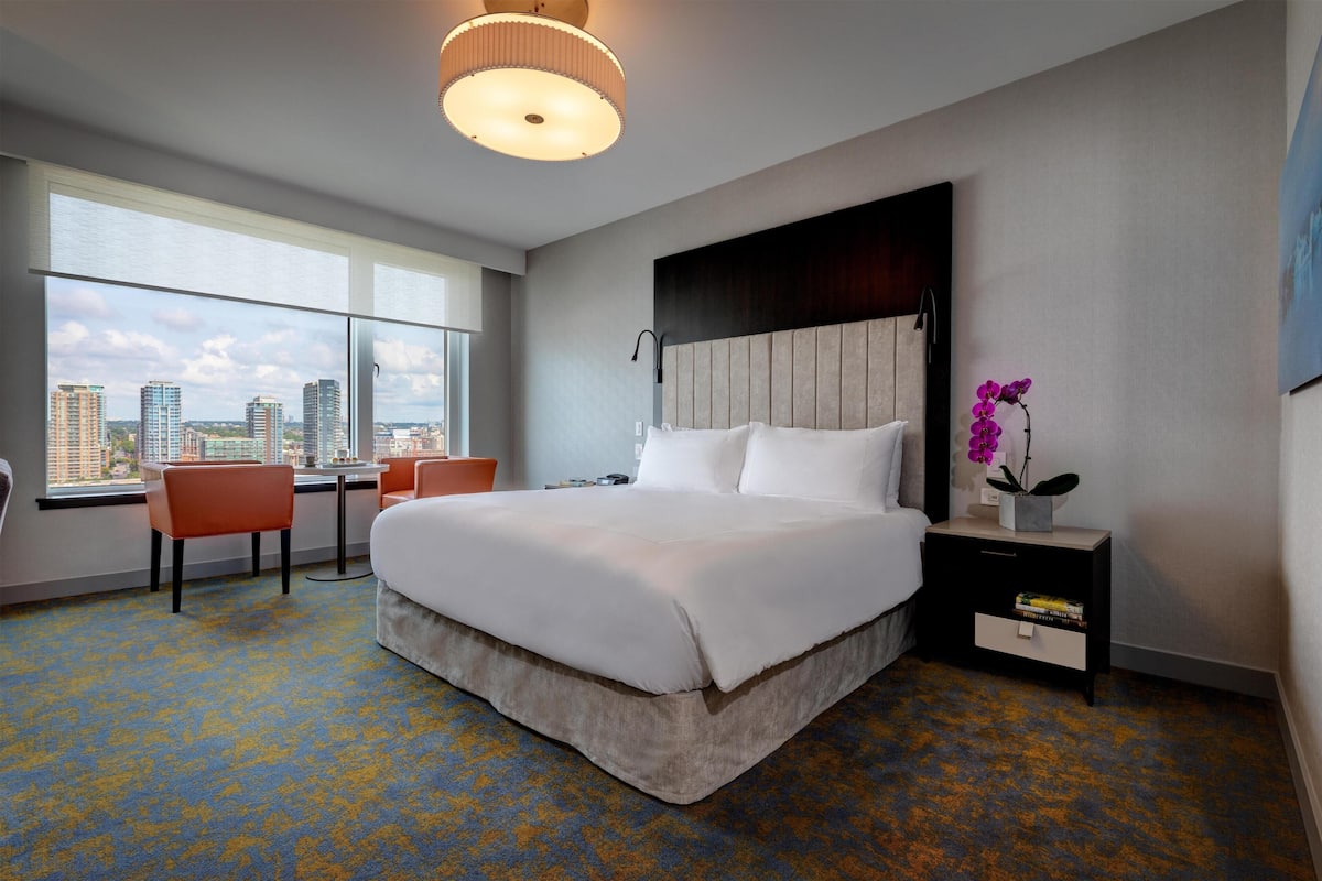 A hotel room with a large bed, a desk, and a chair, a nightstand with a vase of flowers, and a city view through large windows.
