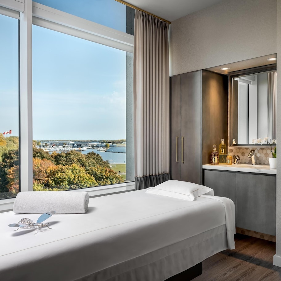A hotel room with a large window, a bed, a small table, a mirror, and a view of a body of water and trees.