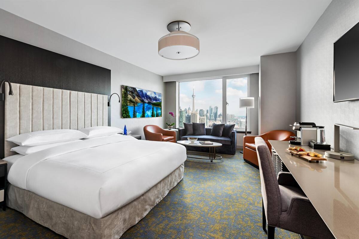 A hotel room with a large bed, a sofa, a dining table, and a city view.