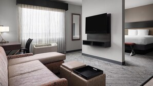 Quarto de hotel com sofá, televisão na parede, cama, escrivaninha com abajur e janela com cortinas.