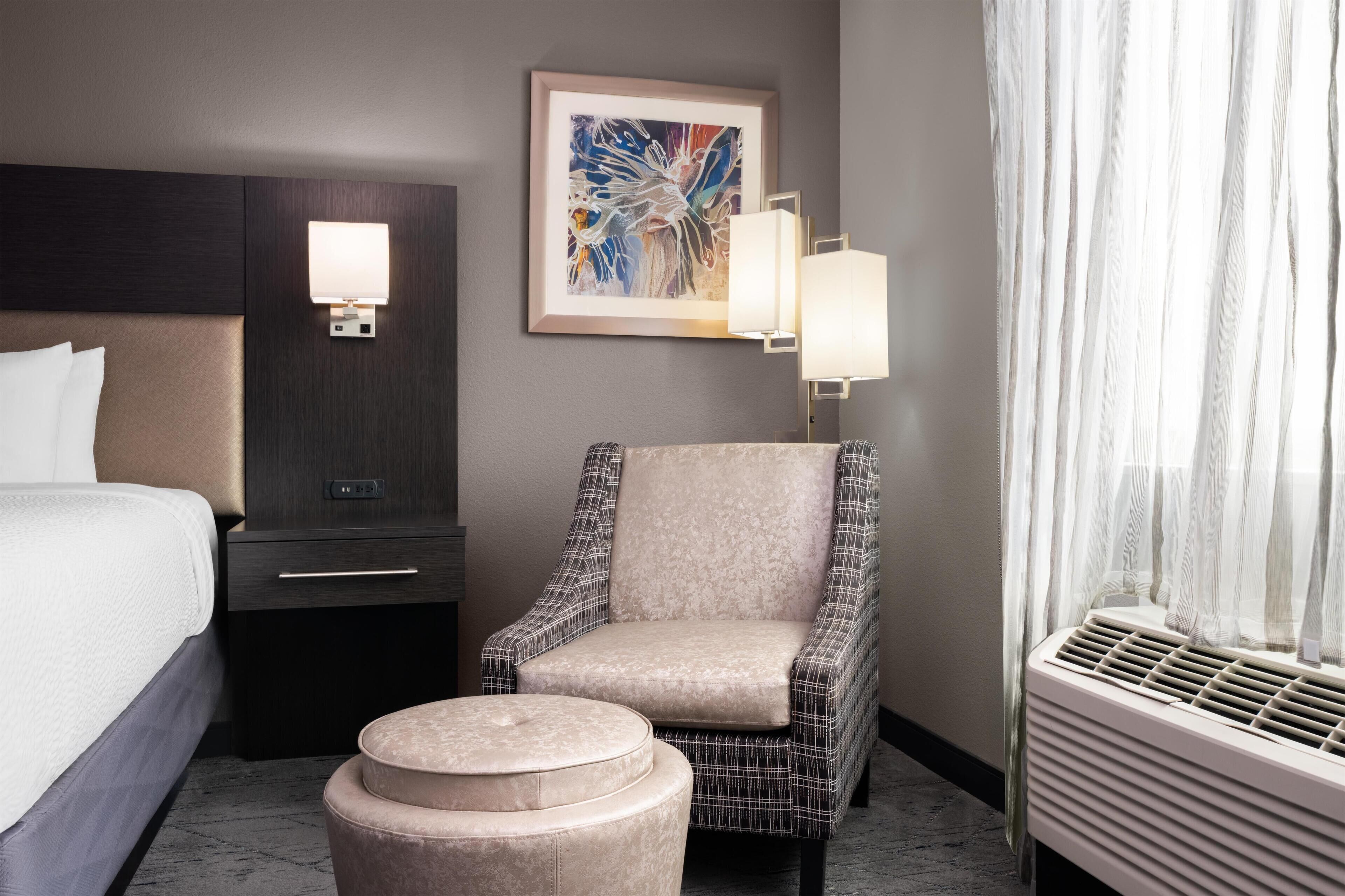Ein Hotelzimmer mit Bett, Stuhl, Nachttisch, Lampe, einem Gemälde und einem Fenster mit Vorhängen.