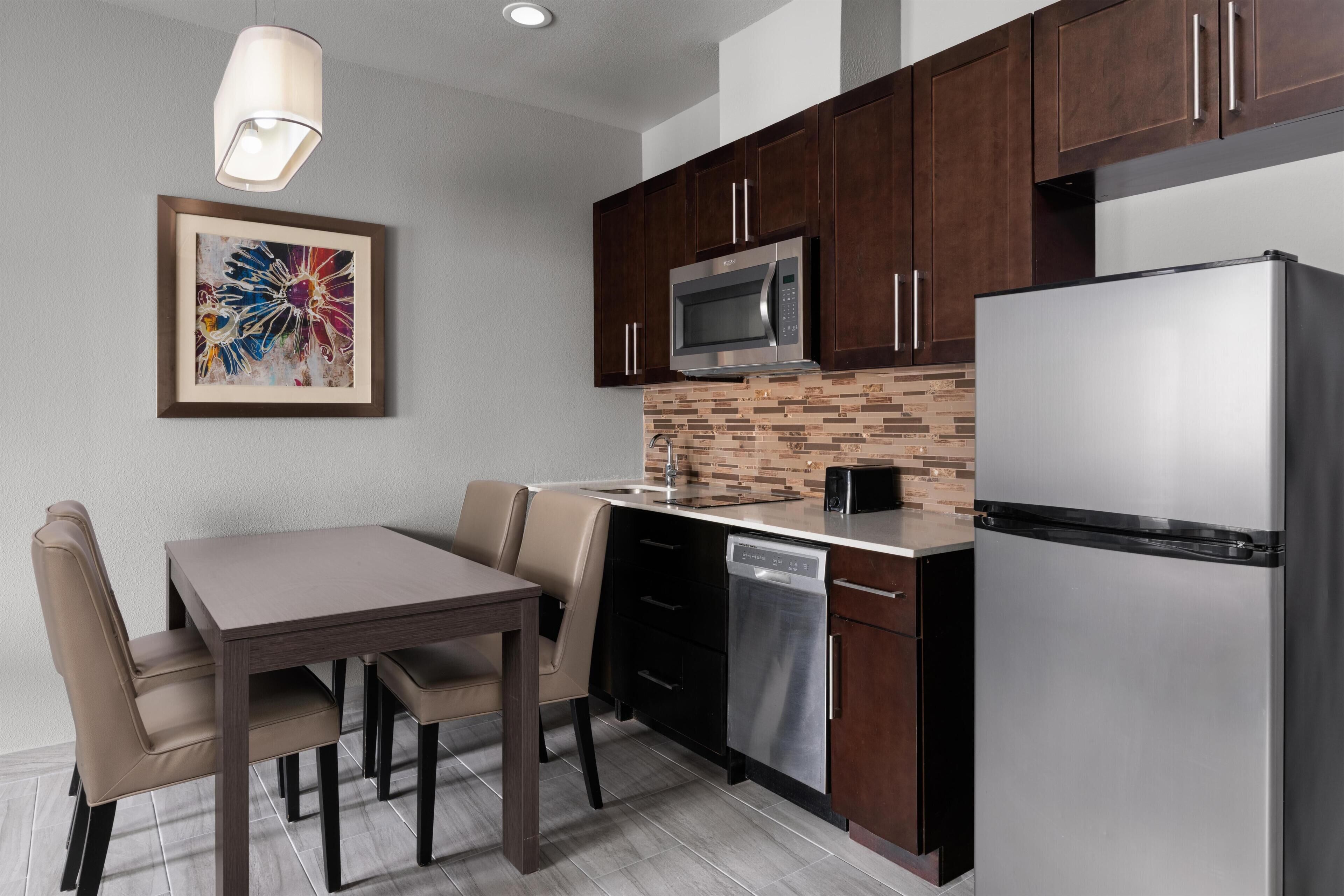 A modern kitchen with dark wooden cabinets, a stainless steel refrigerator, a microwave, and a dishwasher. There is a dining table with chairs and a pendant light above it.