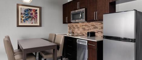 A modern kitchen with dark wooden cabinets, a stainless steel refrigerator, a microwave, and a dishwasher. There is a dining table with chairs and a pendant light above it.