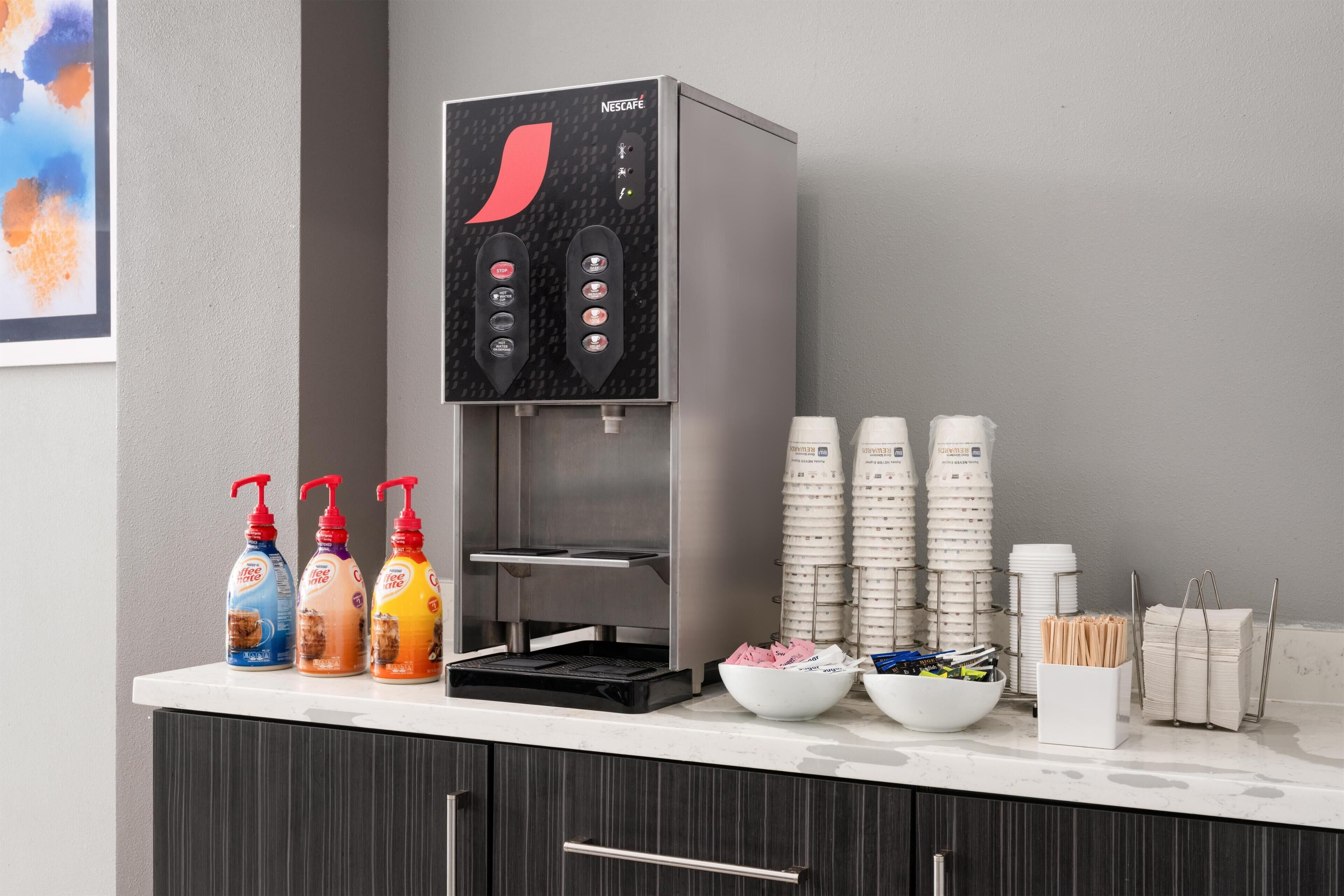 Nescafé-kahvinkeitin, kertakäyttöisiä kahvikuppeja ja kulho, jossa on aterimia, ovat tasolla.
