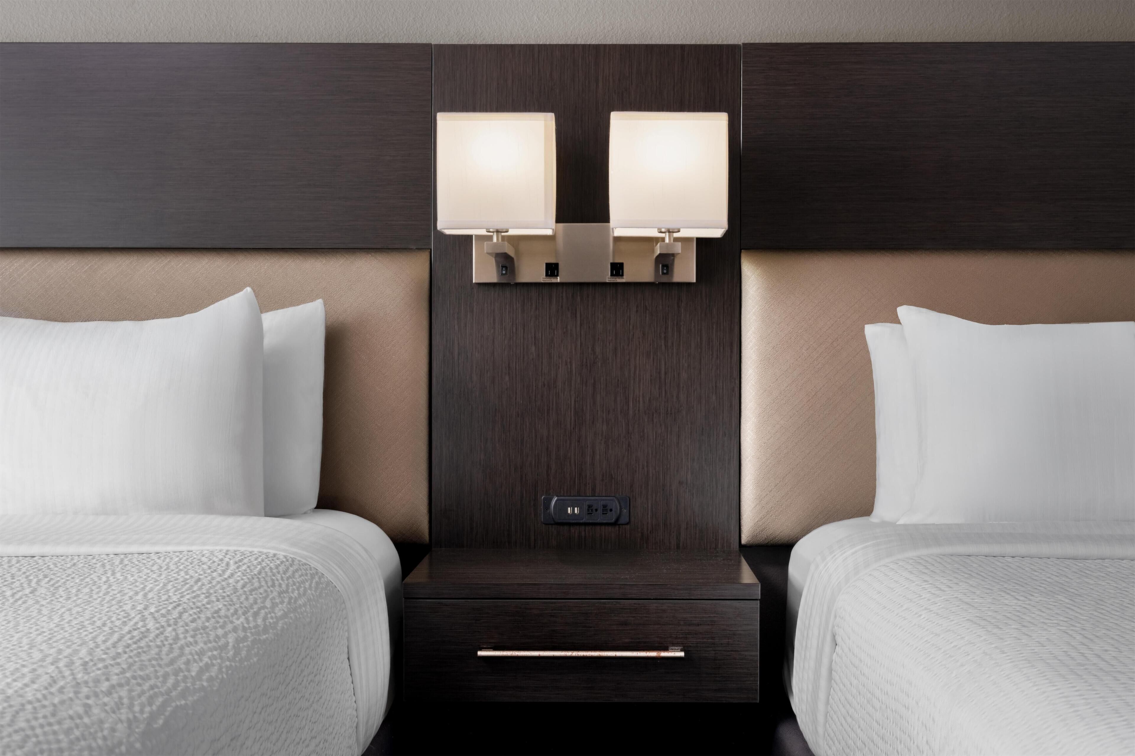 A hotel room with two beds, a dark wood headboard, bedside lamps, a control panel, and a drawer.