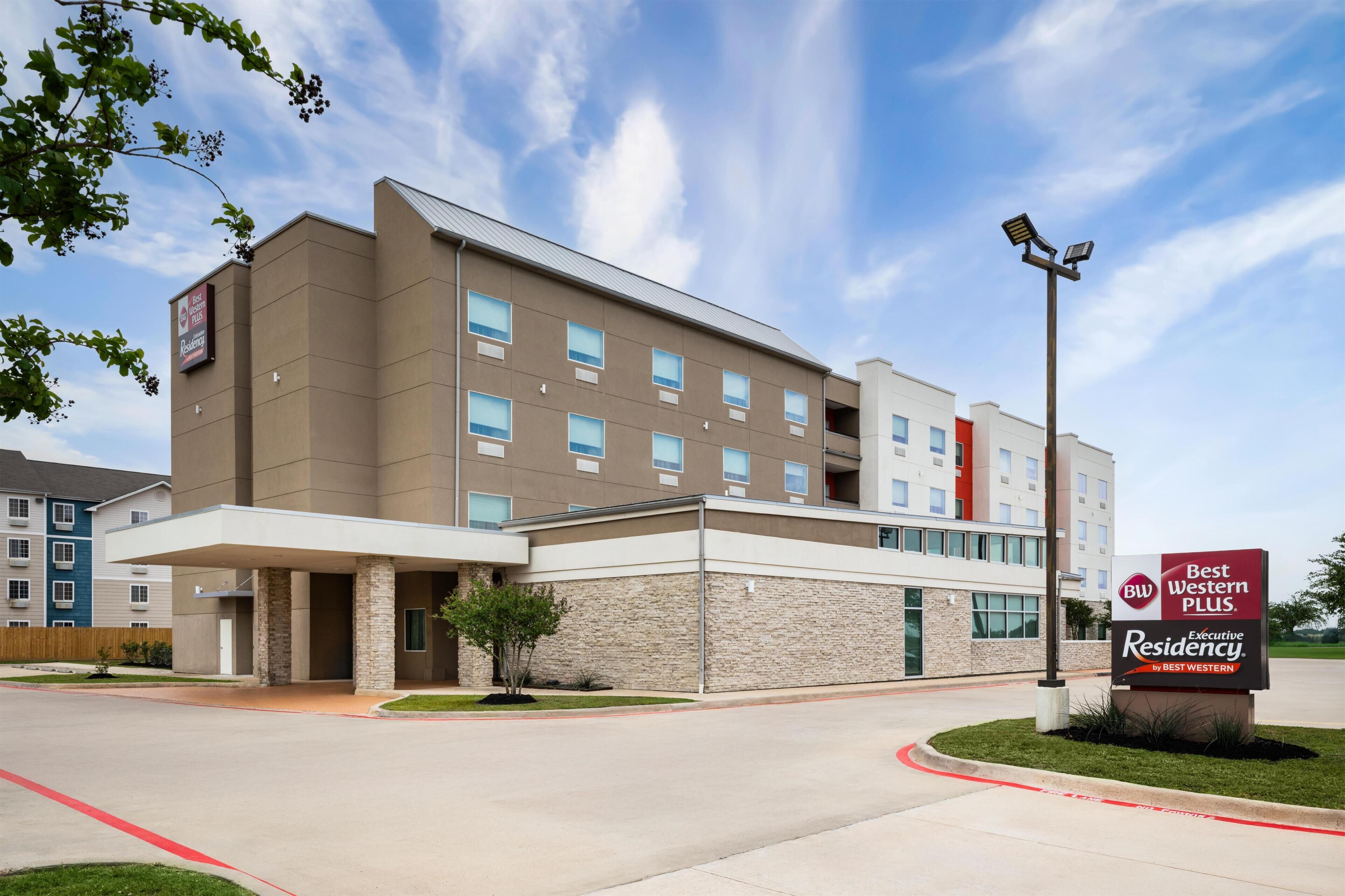 A multi-story hotel with a modern design, featuring a prominent entrance, a landscaped area with a small tree, and a sign indicating it is a Best Western Plus Executive Residency.
