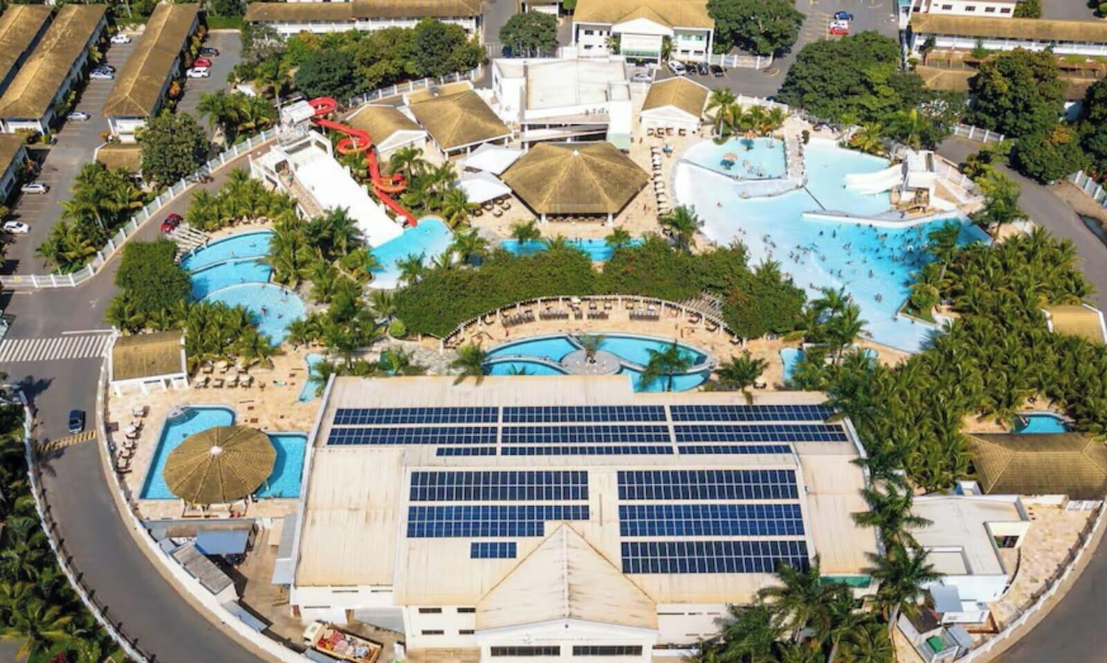 Vista aérea de un complejo turístico con múltiples piscinas, un edificio grande con paneles solares y una carretera con coches estacionados.