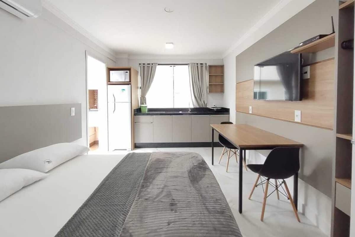 Ett modernt sovrum med en säng, ett nattduksbord och ett skrivbord med en stol. Det finns också en liten köksdel med ett kylskåp och ett fönster med gardiner.