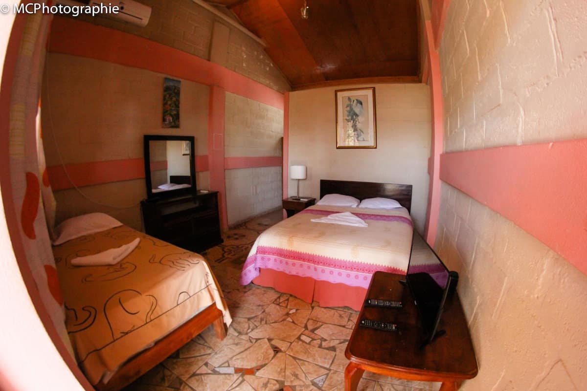 A hotel room with two beds, a nightstand, a bedside table, and a wall-mounted lamp.