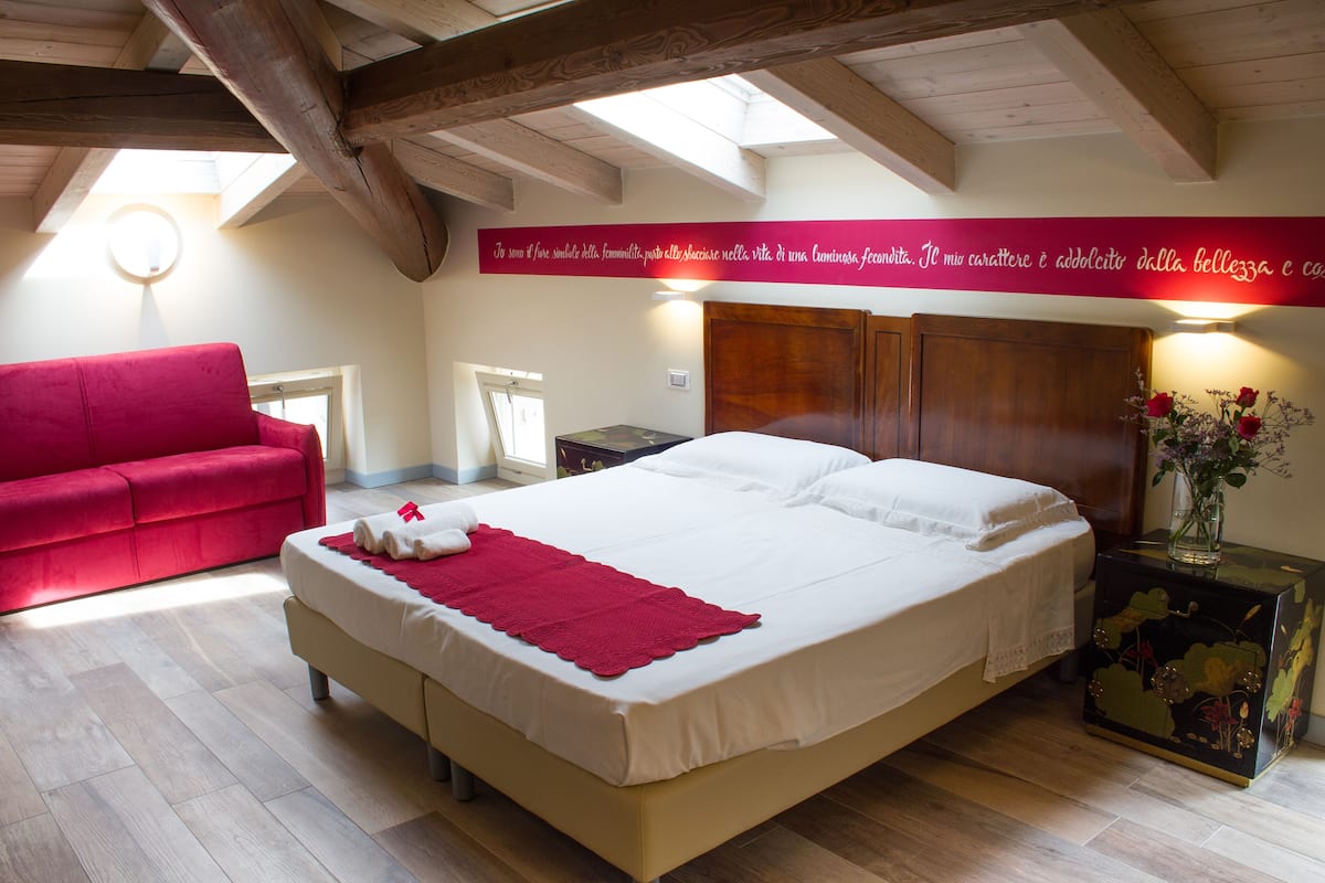A bedroom with a large bed, a red sofa, wooden beams on the ceiling, and a wall with a red banner.