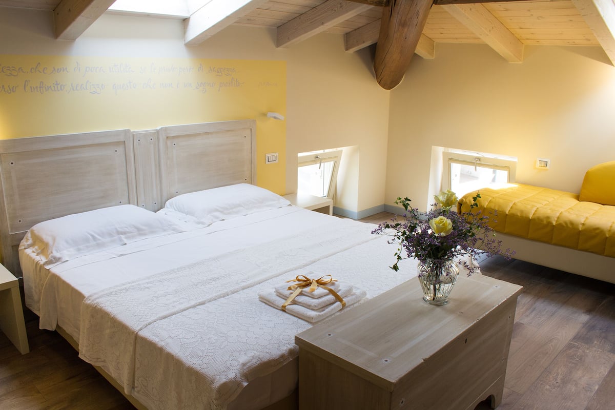 A bedroom with a large bed, a bedside table with a vase of flowers, and a window with a view of the outside.