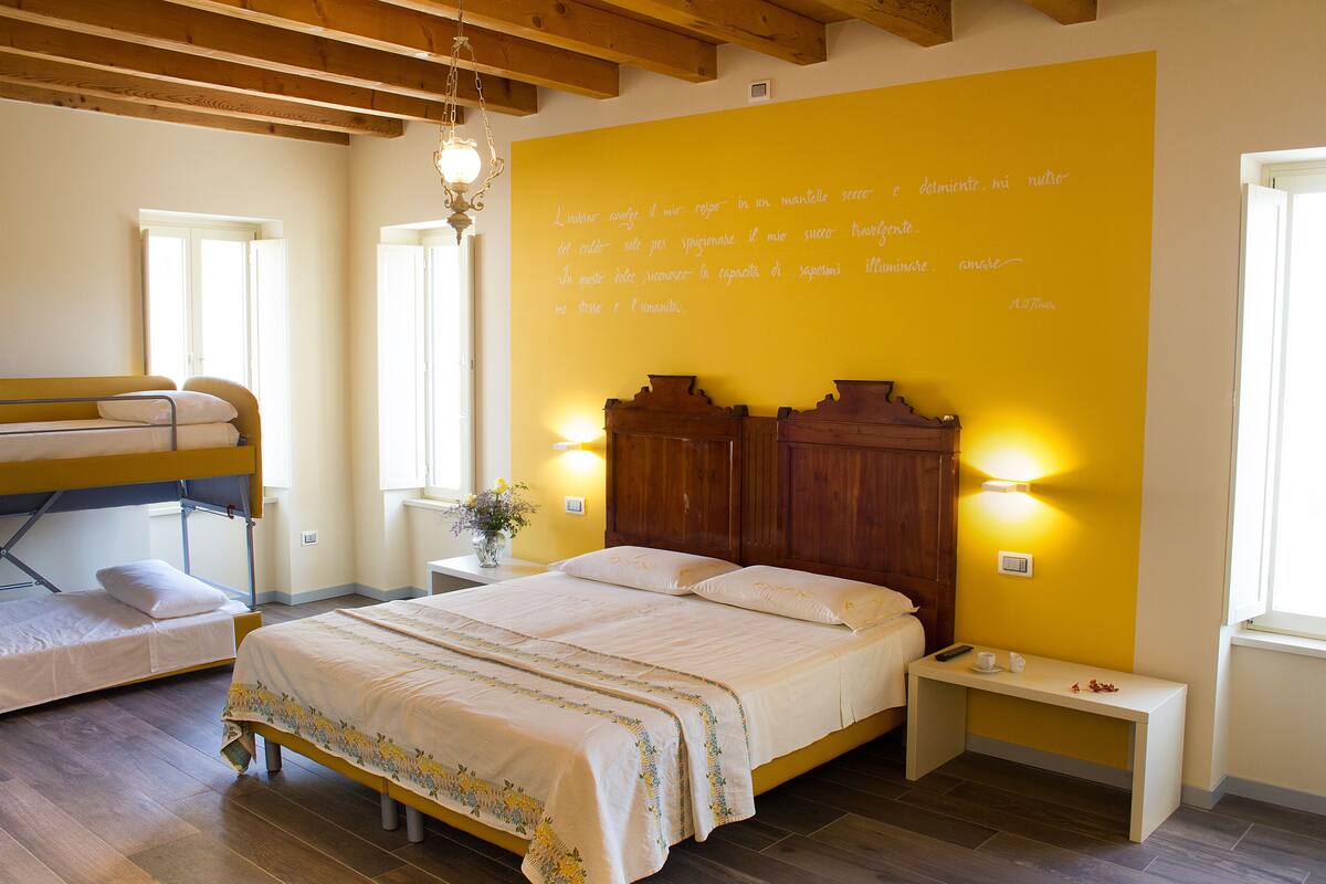 A bedroom with a bunk bed, a single bed, a bedside table with a lamp, and a large yellow wall with text.