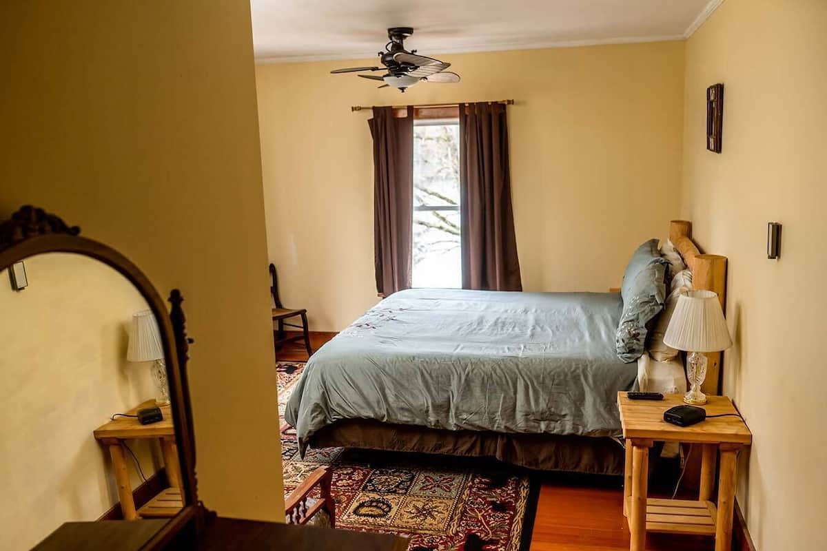 Une chambre à coucher comprenant un lit, une table de chevet en bois, une lampe, un ventilateur de plafond, une fenêtre avec des rideaux et une chaise en bois.
