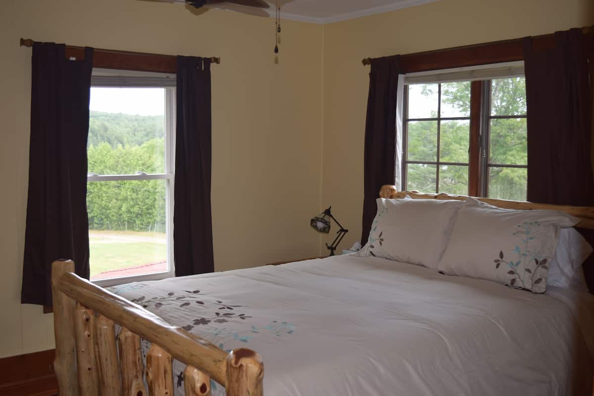 Une chambre à coucher avec un cadre de lit en bois, du linge de lit blanc et deux fenêtres avec des rideaux.