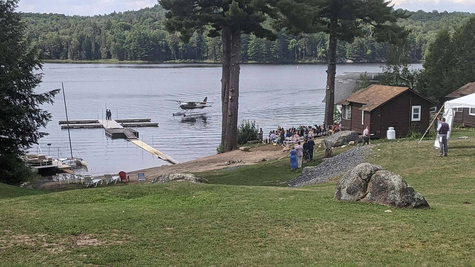 Ett område vid en sjö med en brygga, ett litet flygplan och en grupp människor som samlats nära en byggnad.