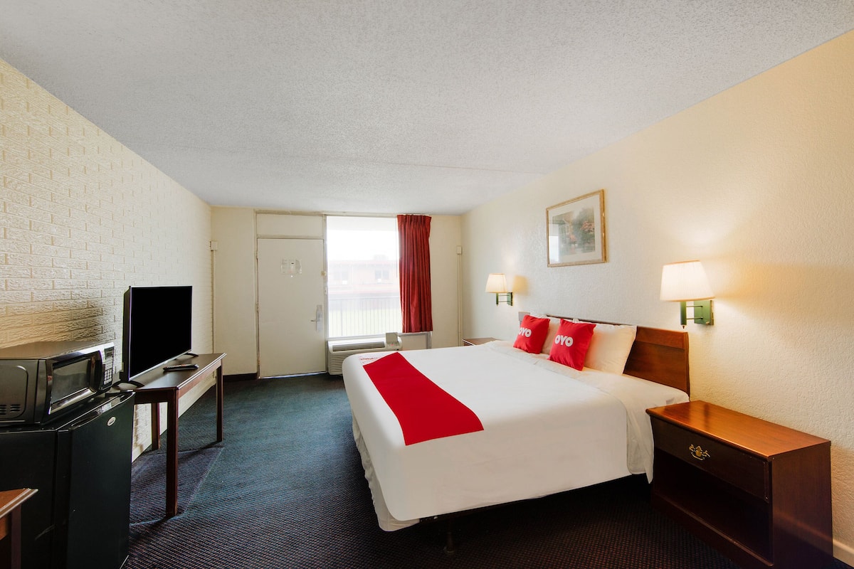 Une chambre d’hôtel comprenant un lit, un bureau avec une télévision et un micro-ondes, une fenêtre avec des rideaux et une table de chevet avec une lampe.