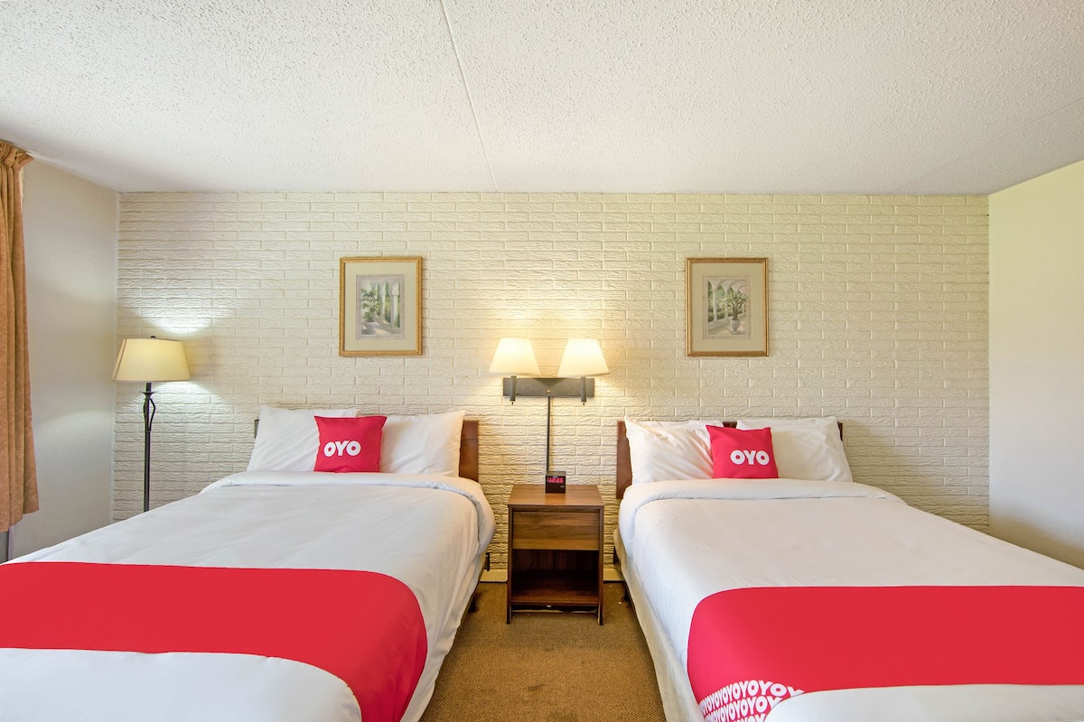 Une chambre d’hôtel avec deux lits, chacun recouvert d’une courtepointe rouge et blanche et agrémenté d’oreillers portant le logo « OYO ». Deux lampes murales sont fixées entre les lits, et des tableaux encadrés ornent le mur en briques situé au-dessus.