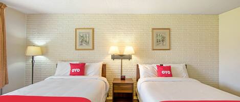 A hotel room with two beds, each with a red and white bedspread and pillows with the "OYO" logo. There are two wall-mounted lamps between the beds, and framed pictures on the brick-patterned wall above.