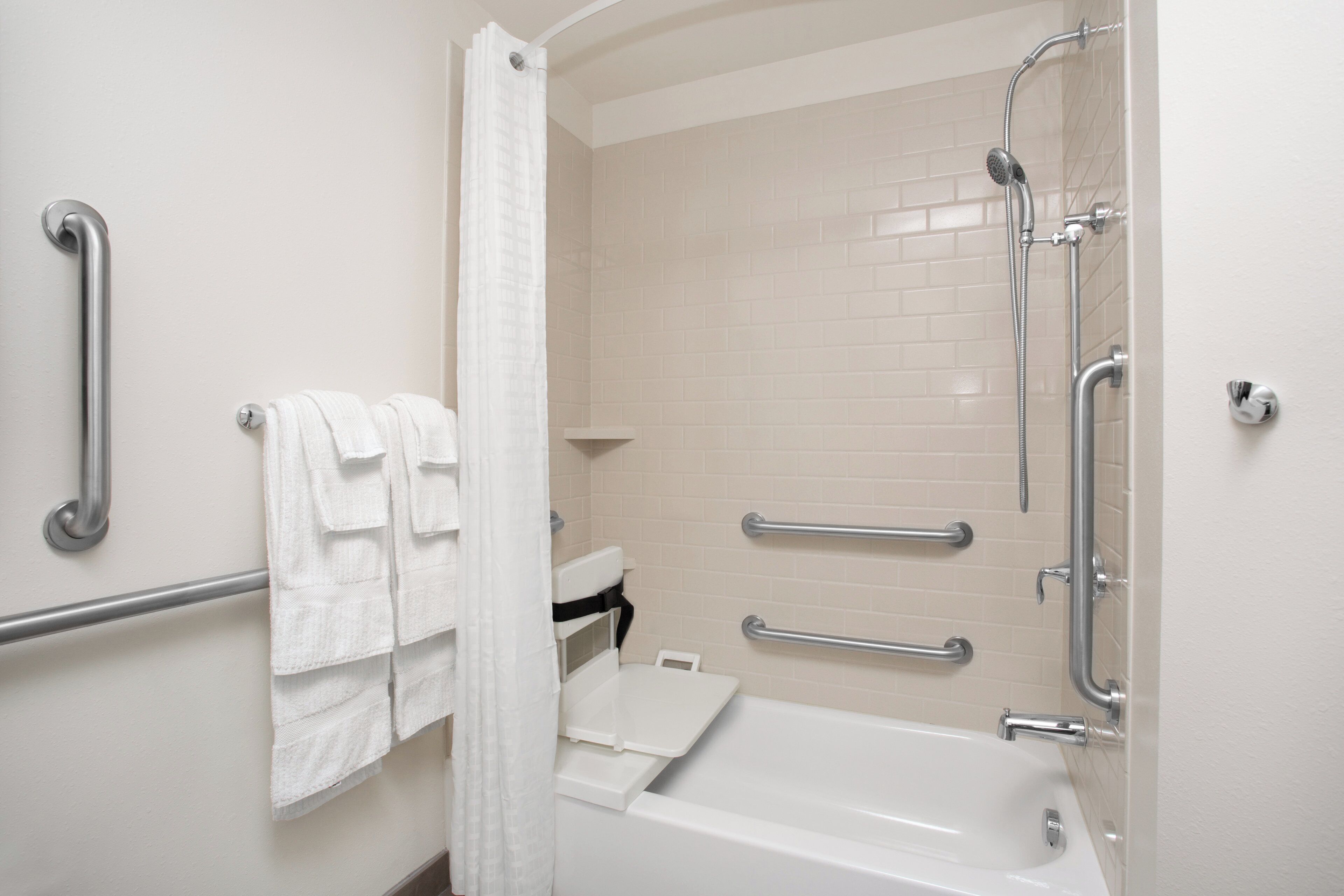 Une salle de bain avec une baignoire blanche, une douche équipée d’un pommeau de douche à main, des barres d’appui et un porte-serviettes blanc.