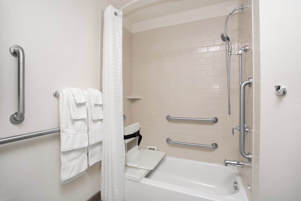 Een badkamer met een witte badkuip, een douche met een handdouche, greepstangen en een witte handdoekhouder.