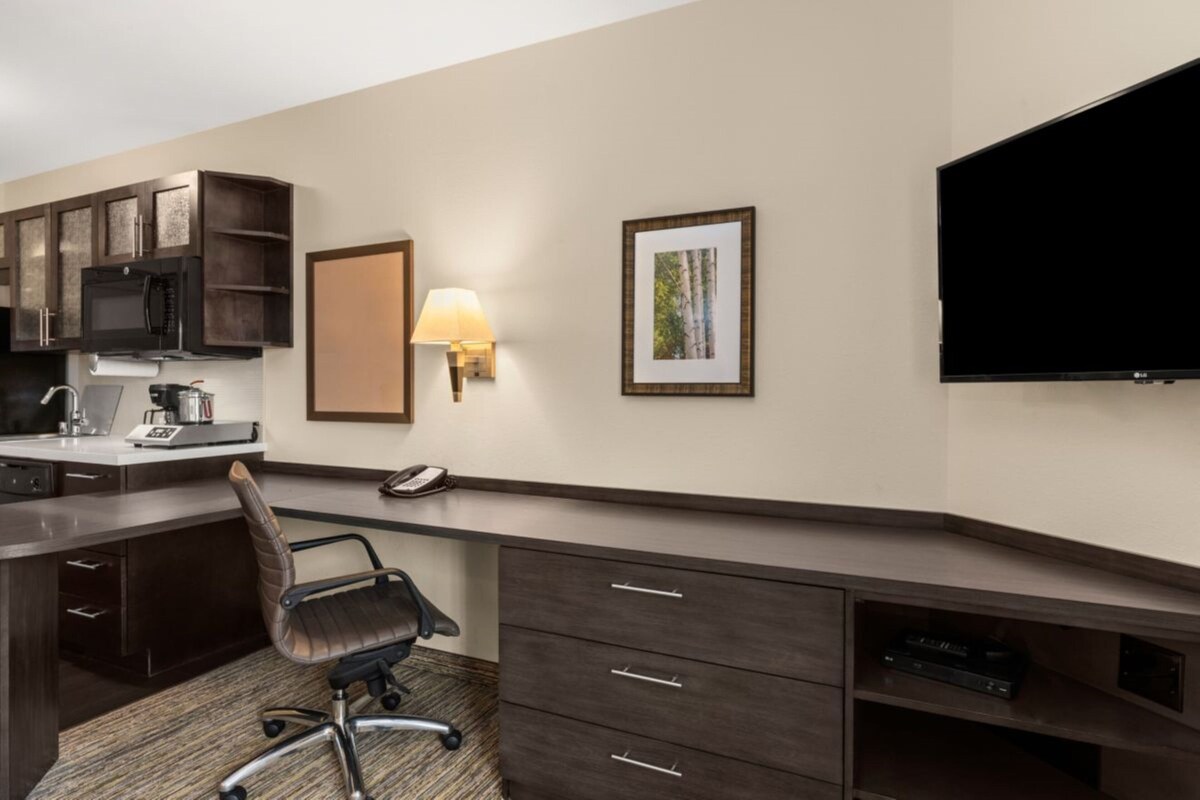 Een hotelkamer met een kitchenette, een bureau met stoel, een flatscreen televisie, een lamp en een ingelijst schilderij aan de muur.
