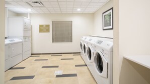 Une buanderie avec un sol carrelé, une machine à laver et un sèche-linge fixés au mur, une bouche d’aération et un panneau indiquant que les machines à laver et les sèche-linge sont à disposition gratuitement.
