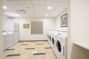 Une buanderie avec un sol carrelé, une machine à laver et un sèche-linge fixés au mur, une bouche d’aération et un panneau indiquant que les machines à laver et les sèche-linge sont à disposition gratuitement.