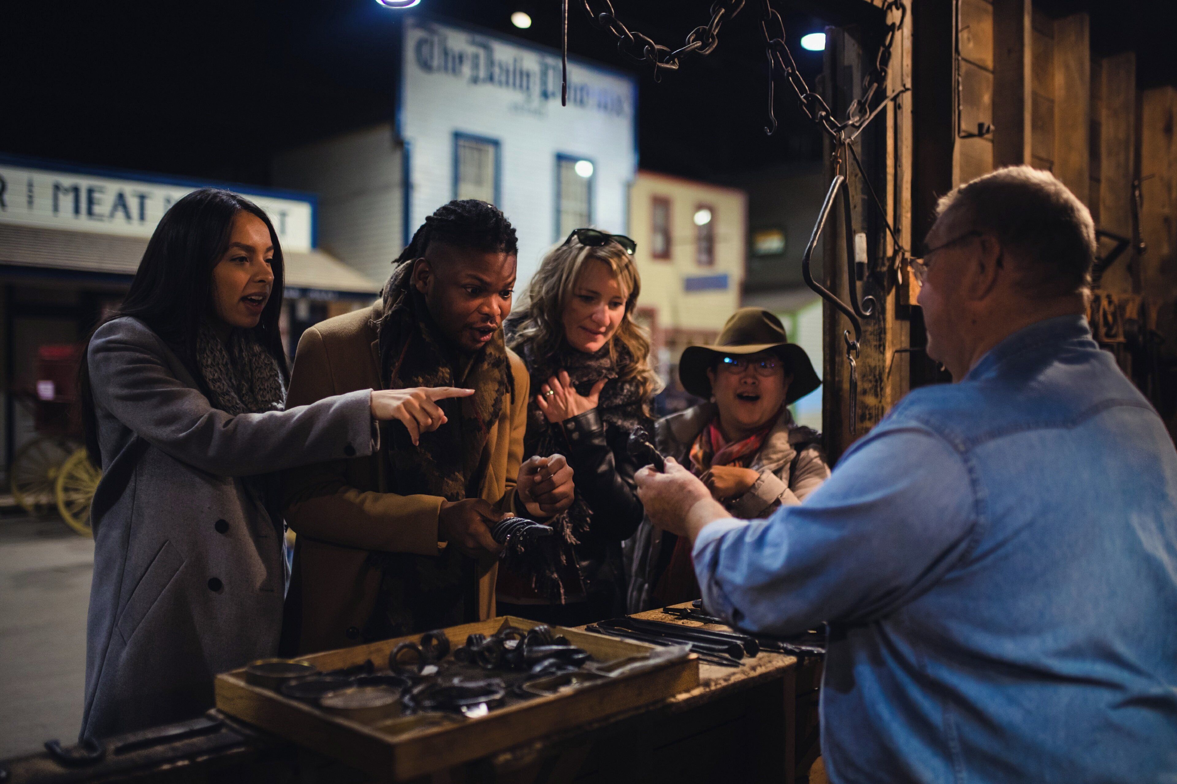A group of people gathered around a table with various items, possibly for sale, in an outdoor setting with buildings in the background.