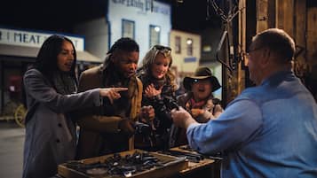 A group of people gathered around a table with various items, possibly for sale, in an outdoor setting with buildings in the background.