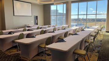 Un salón de conferencias con largas mesas, sillas, una pantalla de proyector y ventanas amplias con vistas.
