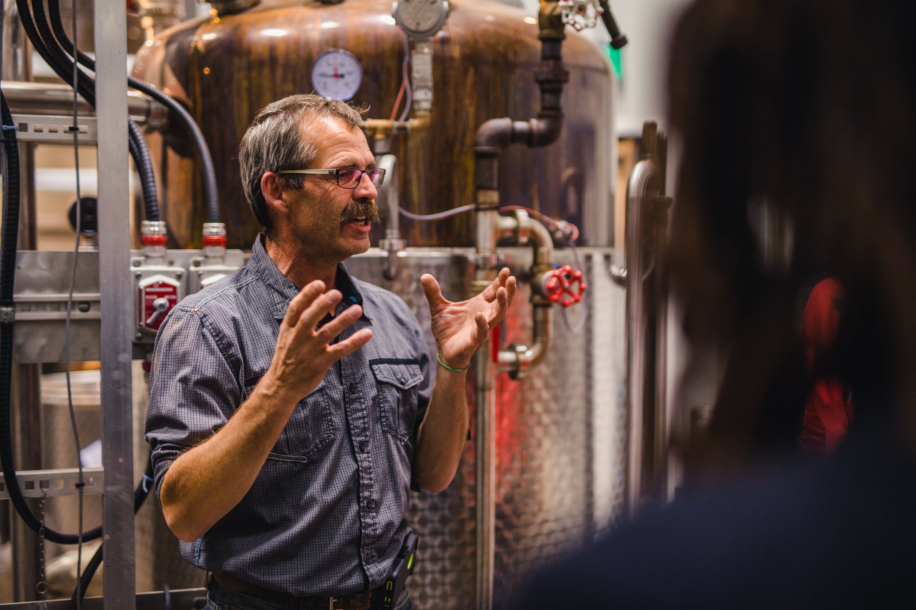 A man in a checkered shirt standing in front of industrial equipment, likely in a brewery or distillery.
