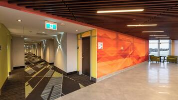 A hotel corridor with a geometric patterned carpet, a wall with directional signage, and a seating area with chairs.