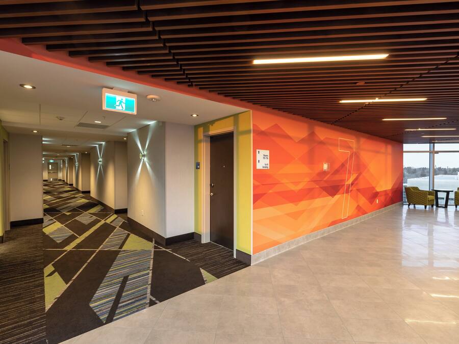 A hotel corridor with a geometric patterned carpet, a wall with directional signage, and a seating area with chairs.
