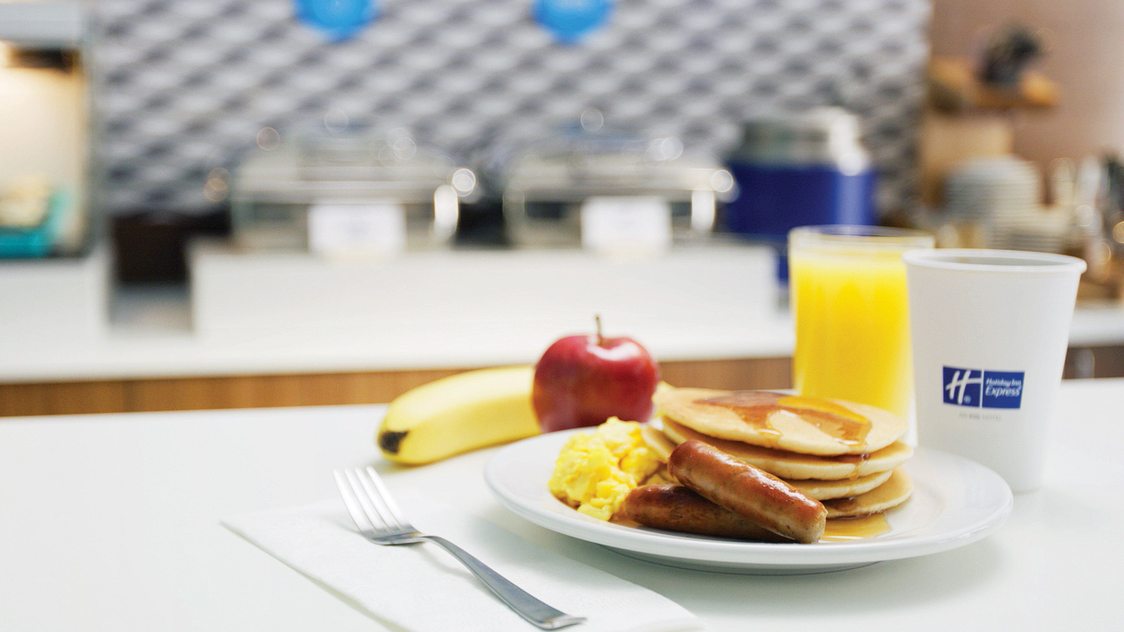 A breakfast plate with pancakes, sausage, and scrambled eggs, an apple, a banana, and a glass of orange juice on a hotel table.