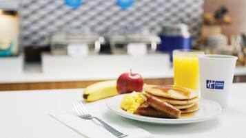 A breakfast plate with pancakes, sausage, and scrambled eggs, an apple, a banana, and a glass of orange juice on a hotel table.