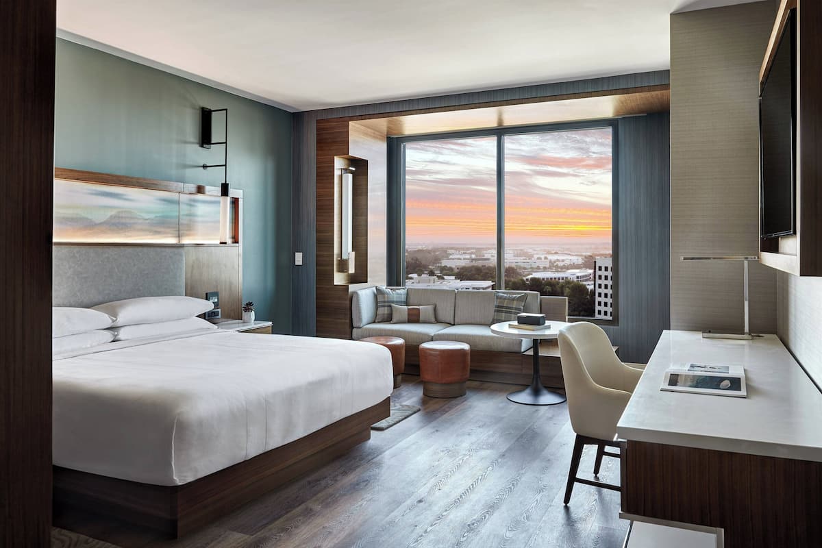 A modern hotel room with a large bed, a seating area with a view of the sunset, and a desk with a chair.