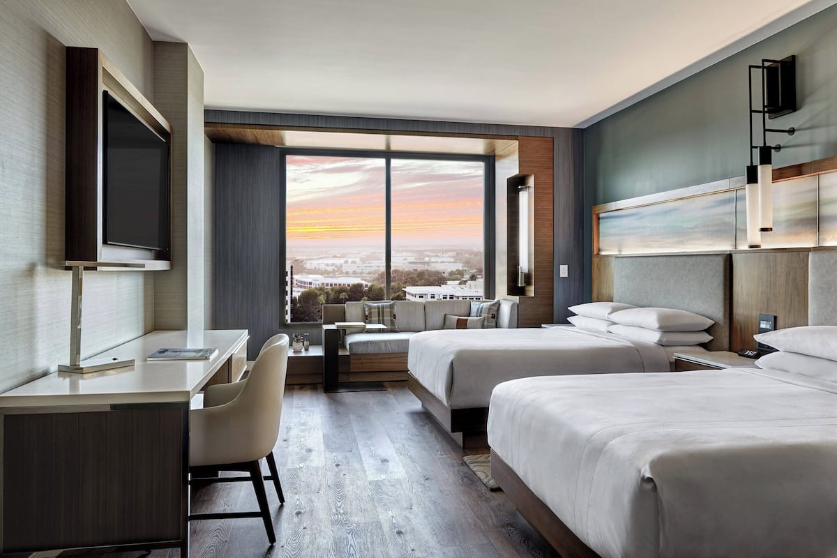 A hotel room with a large window, a desk, two beds, and a TV.