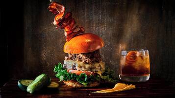 A burger with a sausage on a stick, a glass of iced tea, and some vegetables on a wooden table.